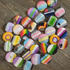 May include: A collection of colourful round buttons with various designs, including rainbow stripes, hearts, and peace signs. The buttons are made of metal and have a shiny finish.