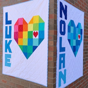 May include: Two white fabric squares with blue and white trim. Each square has a large heart made of colorful squares. One heart is rainbow colored and the other is blue. The squares have the names "LEKE" and "NOLAN" written vertically in blue.
