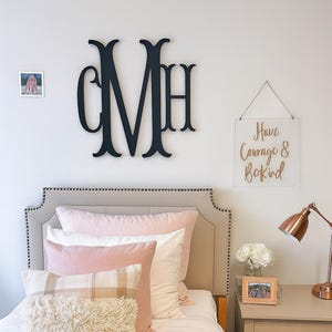 May include: A bedroom scene featuring a bed with a beige headboard and various pillows in shades of pink and white. A decorative wooden monogram is mounted on the wall above the bed. A sign with the words "Have Courage & Be Kind" hangs on the wall.