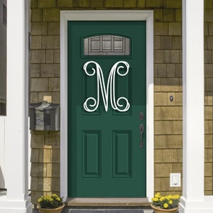 May include: A white, decorative letter "M" is mounted on a dark green front door. The door has a glass panel at the top and is framed by white trim. The house exterior features a mailbox and a shingled wall.