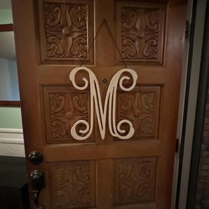 May include: A white wooden letter "M" hanging on a wooden door. The letter has a decorative, cursive design. The door is a medium brown color and has carved panels. A black string is used to hang the letter.