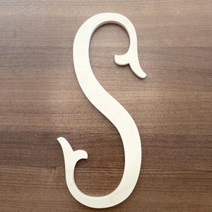 May include: A large, unpainted wooden letter "S" with a decorative, curved design. The letter is light in color and stands out against the dark brown wood grain background. The "S" has a classic, elegant style.