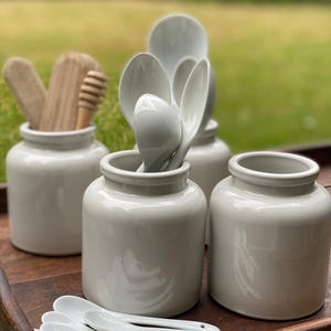 Puede incluir: Una colección de utensilios de cocina y tarros de almacenamiento. La imagen presenta cuatro tarros de cerámica gris claro, algunos llenos de cucharas blancas y utensilios de madera. Una bandeja de madera contiene los tarros y cucharas blancas adicionales.