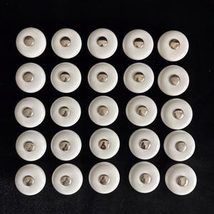 May include: Twenty-four white ceramic cabinet knobs with a silver-colored metal center. The knobs are arranged in a grid pattern against a black background. These knobs are suitable for furniture restoration or new builds.