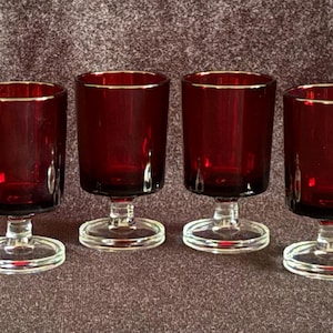 May include: A set of four matching drinking glasses featuring deep red, translucent glass bowls and clear glass stems with circular bases. Each glass has a straight-sided cylindrical shape and a smooth, polished finish throughout the design.
