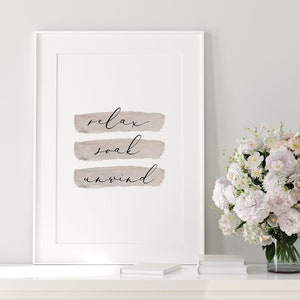 May include: Framed wall art with the words "relax, soak, unwind" in elegant script over taupe brushstrokes. The artwork is displayed on a white surface, with a vase of white flowers and books in the background.