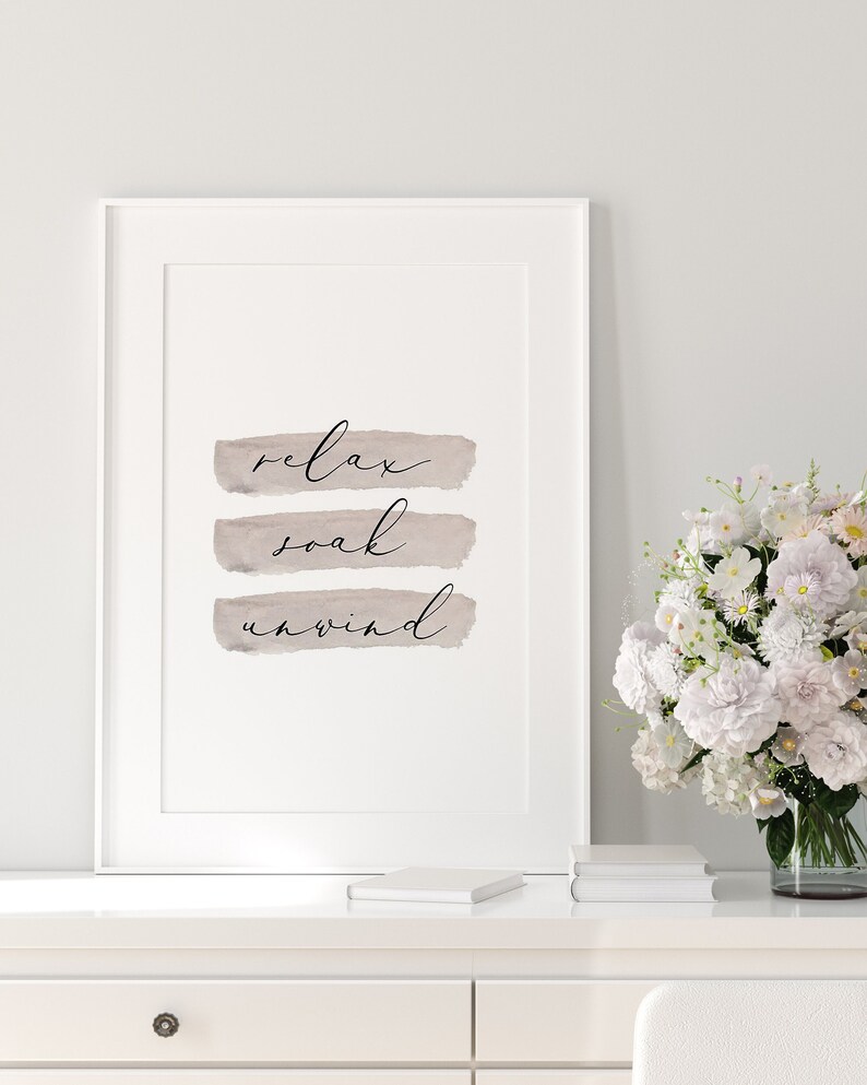 May include: Framed wall art with the words "relax, soak, unwind" in elegant script over taupe brushstrokes. The artwork is displayed on a white surface, with a vase of white flowers and books in the background.