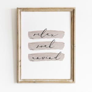 May include: A framed print with the words "relax, soak, unwind" in elegant script. The words are layered over three horizontal, muted gray brushstrokes. The frame is a light, distressed gold color, adding a touch of rustic charm to the artwork.