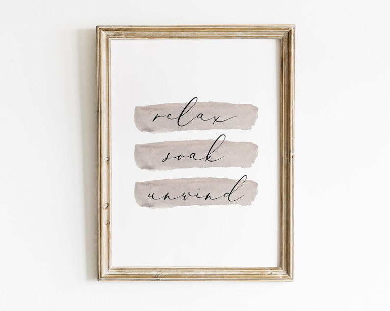 May include: A framed print with the words "relax, soak, unwind" in elegant script. The words are layered over three horizontal, muted gray brushstrokes. The frame is a light, distressed gold color, adding a touch of rustic charm to the artwork.