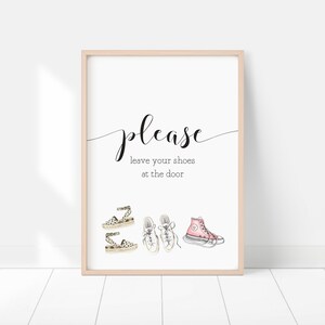 Op de afbeelding: Een wit bord met zwarte tekst die zegt "Alstublieft laat je schoenen bij de deur". Het bord is ingelijst in een licht houten frame. Er zijn vier tekeningen van schoenen onder de tekst: twee paar sandalen en een paar sneakers.