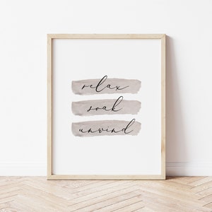 May include: A framed print with the words "relax, soak, unwind" in black script over three horizontal, muted taupe brushstrokes. The frame is a light wood color, and the background is white.