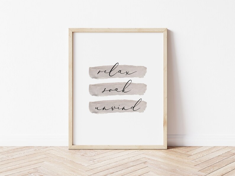 May include: A framed print with the words "relax, soak, unwind" in black script over three horizontal, muted taupe brushstrokes. The frame is a light wood color, and the background is white.