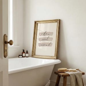 May include: A framed print with the words "relax, soak, unwind" in elegant script. The gold frame surrounds a cream-colored background with three horizontal beige brushstrokes. The print is displayed in a bathroom setting, leaning against a white bathtub.