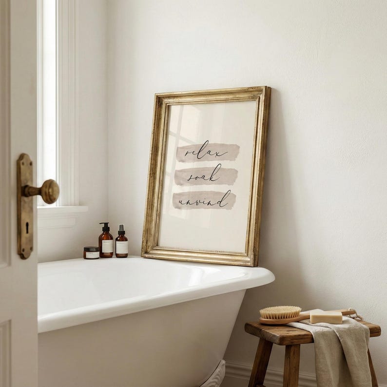 May include: A framed print with the words "relax, soak, unwind" in elegant script. The gold frame surrounds a cream-colored background with three horizontal beige brushstrokes. The print is displayed in a bathroom setting, leaning against a white bathtub.