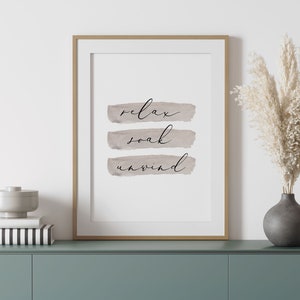 May include: Framed art print with the words "relax, soak, unwind" in elegant script over three horizontal watercolor-style brushstrokes. The print is in a gold frame, displayed on a teal cabinet with decorative items.