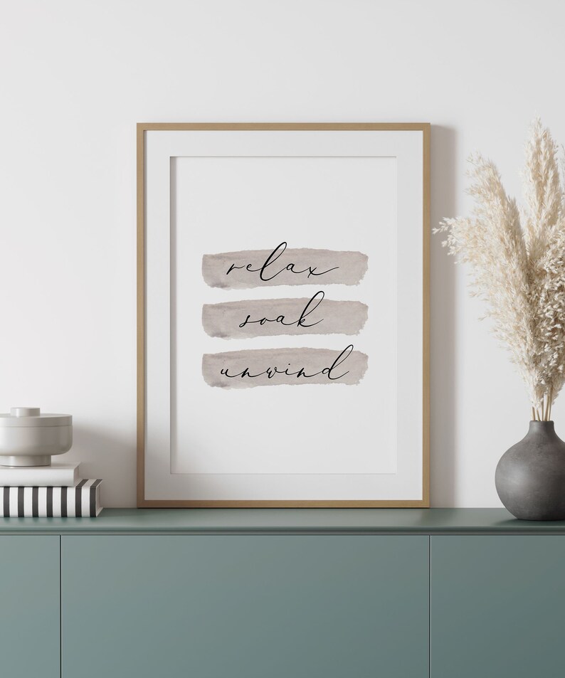 May include: Framed art print with the words "relax, soak, unwind" in elegant script over three horizontal watercolor-style brushstrokes. The print is in a gold frame, displayed on a teal cabinet with decorative items.