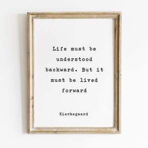May include: A framed print with a light brown wooden frame. The print features the quote "Life must be understood backward. But it must be lived forward" attributed to Kierkegaard, in a typewriter-style font on a white background.