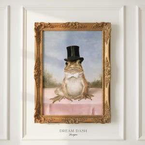 May include: A framed wall art print featuring a painting of a frog wearing a black top hat and a white bow tie. The frog sits on a pink surface against a light blue sky background. The artwork is enclosed in an ornate, gold-coloured frame.