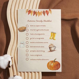 Op de afbeelding: Een beige Autumn Family Checklist met illustraties van herfstactiviteiten. De lijst bevat het bakken van een appeltaart, het kijken van een Halloween-film en het decoreren van pompoenen. Boven hangt een slinger van herfstbladeren.