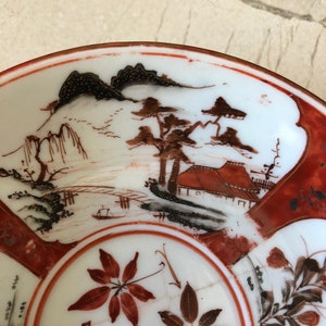 May include: A white porcelain bowl with a red rim and a hand-painted design. The design features a landscape with mountains, trees, and a house. The bowl also has a floral design with red and gold leaves.
