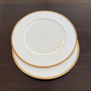 May include: Two white ceramic plates with a gold rim are stacked on a dark brown wooden surface. The plates are round and have a simple, elegant design. The gold rim adds a touch of sophistication to the plates.