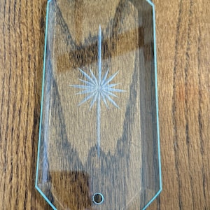 May include: Clear glass decorative tray with a geometric shape and a starburst design. The tray has a light blue border and a small hole at the bottom. The tray is on a wooden surface.