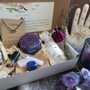 May include: A gift box filled with items for a Libra zodiac sign. The box contains a white sage smudge stick, a small bottle of essential oil, a crystal, a candle, a small tin with a Libra symbol, and a necklace with a Libra symbol. The box is decorated with a floral design and the words "Libra" and "September 23 - October 22".