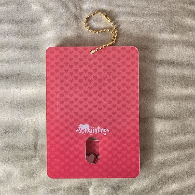 May include: A rectangular pink card with a heart pattern and a gold-colored chain. The card has a small cutout revealing a heart design. The text "Yokaiihime" is printed on the card.