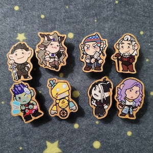 Op de afbeelding: Acht houten pinnen met verschillende cartoonfiguren. De figuren omvatten een robot, een persoon met een zwaard, een persoon met een staf, een persoon met een kroon, een persoon met een hoed, een persoon met een baard, een persoon met een boog en pijl en een persoon met een zwaard.