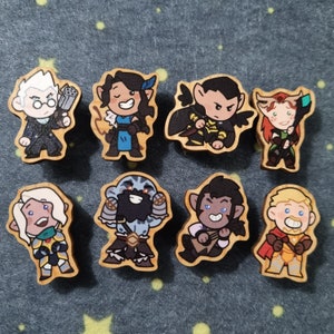 May include: Eight wooden character pins with different designs. The characters are all cartoon-style and have different facial expressions. The pins are painted in various colors, including blue, green, red, yellow, black, and white.