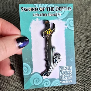Puede incluir: Un pin de esmalte negro que representa una espada con un ojo amarillo y un diseño de ola azul. El pin está en una tarjeta que dice "Sword of the Depths" y "Critical Role Enamel Pin".
