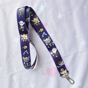 May include: Purple lanyard with a repeating pattern of cartoon characters. The characters are all different and have different expressions. The lanyard has a metal clasp.