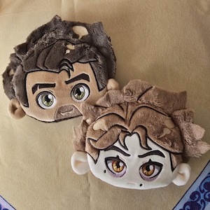 Könnte beinhalten: Zwei Plüschkissen in Form von Cartoon-Gesichtern. Eines hat braunes Haar und einen Bart mit grünen Augen. Das andere hat hellbraunes Haar und goldene Augen. Beide haben detaillierte Gesichtszüge und sind aus weichem, strukturiertem Stoff.