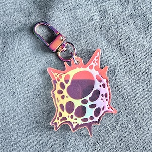 May include: A holographic keychain with a unique, abstract design. The keychain features a colorful, iridescent charm with a series of holes and a jagged outline. It is attached to a metallic, rainbow-colored clasp.
