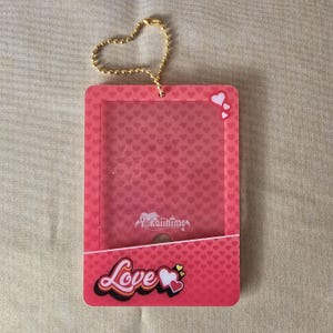 May include: A pink ID badge holder with a heart-patterned background and the word "Love" in a colorful, cursive font. The holder has a clear window and a gold-colored chain.