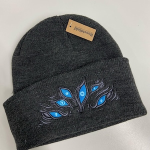 May include: A dark gray beanie with a blue and gray embroidered design of multiple eyes. The beanie has a brown tag with the text "Beechfield Original Wearer".