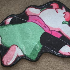 Puede incluir: Una alfombra de dibujos animados con un diseño de personaje en blanco, rosa, verde y negro. La alfombra tiene un borde negro y una forma irregular. El personaje está acostado, con los pies y la cabeza visibles.