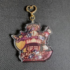 Puede incluir: Un llavero de acrílico transparente con una ilustración de dibujos animados de un edificio rojo y amarillo con un dije en forma de corazón dorado. El edificio tiene un letrero que dice "Hazbin Hotel" y presenta varios personajes de dibujos animados.