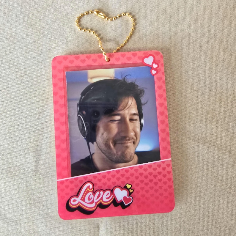 May include: A pink rectangular keychain with a photo of a man wearing headphones. The keychain has a gold heart-shaped chain. The word "Love" is written in a cursive font with hearts.