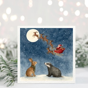 May include: A Christmas card featuring a watercolor illustration of a rabbit and a badger looking up at Santa Claus in his sleigh, pulled by reindeer, flying across a starry night sky. The moon is full and bright.