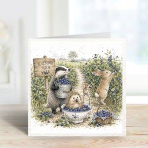 May include: A square greeting card featuring a watercolour illustration of a badger holding a metal bucket of blueberries, a hedgehog in a ceramic bowl, a rabbit picking berries, and a mouse, with a wooden sign reading BLUEBERRY PATCH.