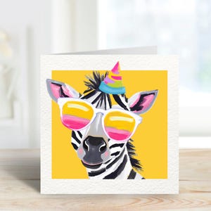 Op de afbeelding: Vierkante wenskaart met een grillige illustratie van een zebra met zonnebril en een feesthoed. De zebra heeft zwart-witte strepen, met roze en gele accenten op de zonnebril en de feesthoed. De achtergrond is heldergeel.