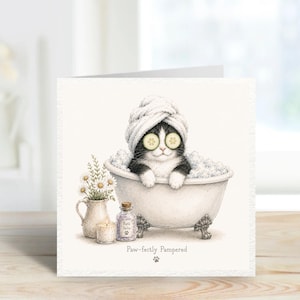 May include: A square greeting card featuring a black and white cat in a clawfoot bathtub with a white towel wrapped around its head and cucumber slices over its eyes. Beside the tub are a vase of daisies, a candle, and bath salts. Text reads Paw-fectly Pampered.