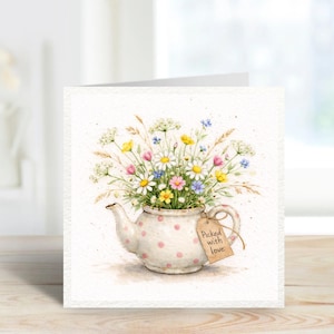 Könnte beinhalten: Aquarell-Grußkarte mit einer Teekanne voller Wildblumen. Die weiße Teekanne hat rosa Tupfen und ein Etikett mit der Aufschrift "Picked with love". Die Blumen umfassen Gänseblümchen, gelbe, rosa und blaue Blüten sowie grünes Laub.