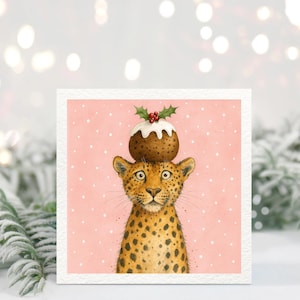 Può includere: Biglietto d'auguri quadrato con l'illustrazione di un leopardo con un budino di Natale in testa. Il leopardo ha un mantello maculato e grandi occhi. Il budino è decorato con glassa bianca, agrifoglio e bacche rosse. Lo sfondo è rosa con punti bianchi.