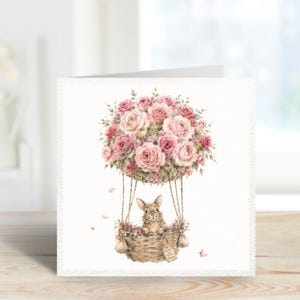 May include: A square greeting card featuring a watercolour illustration of a brown rabbit sitting in a woven basket hot air balloon. The balloon is made of a large bouquet of pink and white roses. A small tag on the basket reads Enjoy the little things.