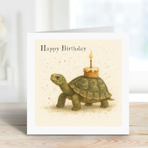 Puede incluir: Tarjeta de cumpleaños con una ilustración en acuarela de una tortuga que lleva un pequeño pastel con una vela encendida. Las palabras "Happy Birthday" están impresas en la parte superior. La tarjeta tiene un borde blanco y un fondo suave y neutro.
