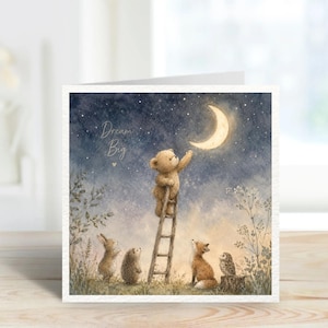 May include: Greeting card with a whimsical illustration. A teddy bear on a ladder reaches for a crescent moon in a starry sky. Other woodland creatures, including a rabbit, hedgehog, fox, and owl, are depicted below. The words "Dream Big" are written on the card.