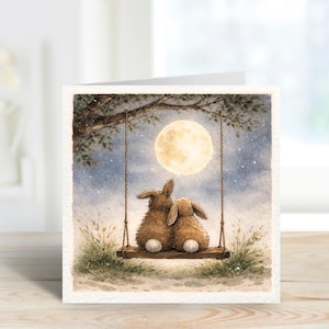 Könnte beinhalten: Eine Grußkarte mit einer Aquarellillustration von zwei braunen Hasen auf einer Schaukel unter einem Vollmond. Der Hintergrund zeigt einen Sternenhimmel und einen Ast. Die Karte hat einen weißen Rand und liegt auf einer Holzoberfläche.
