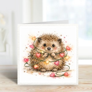 May include: A watercolour illustration of a cute hedgehog with a garland of heart-shaped lights. The hedgehog is brown with a light pink blush and is holding the string of lights. The card is white with a textured finish.
